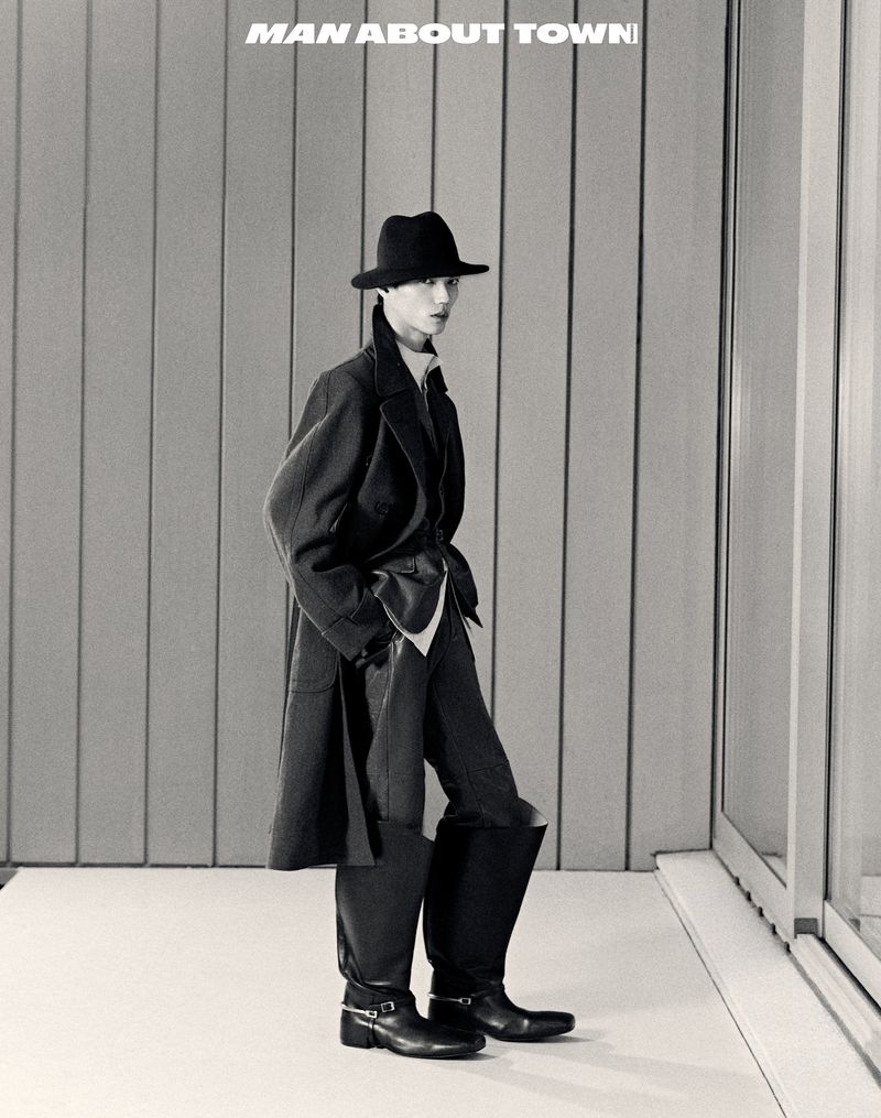Monochrome fashion shot of a person in a dark coat, hat, trousers, and boots, standing in front of a grey wall and white floor.