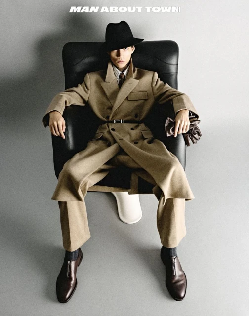 Stylish adult in a double-breasted trench coat, hat, and dress shoes, sitting in a black chair against a neutral studio backdrop.