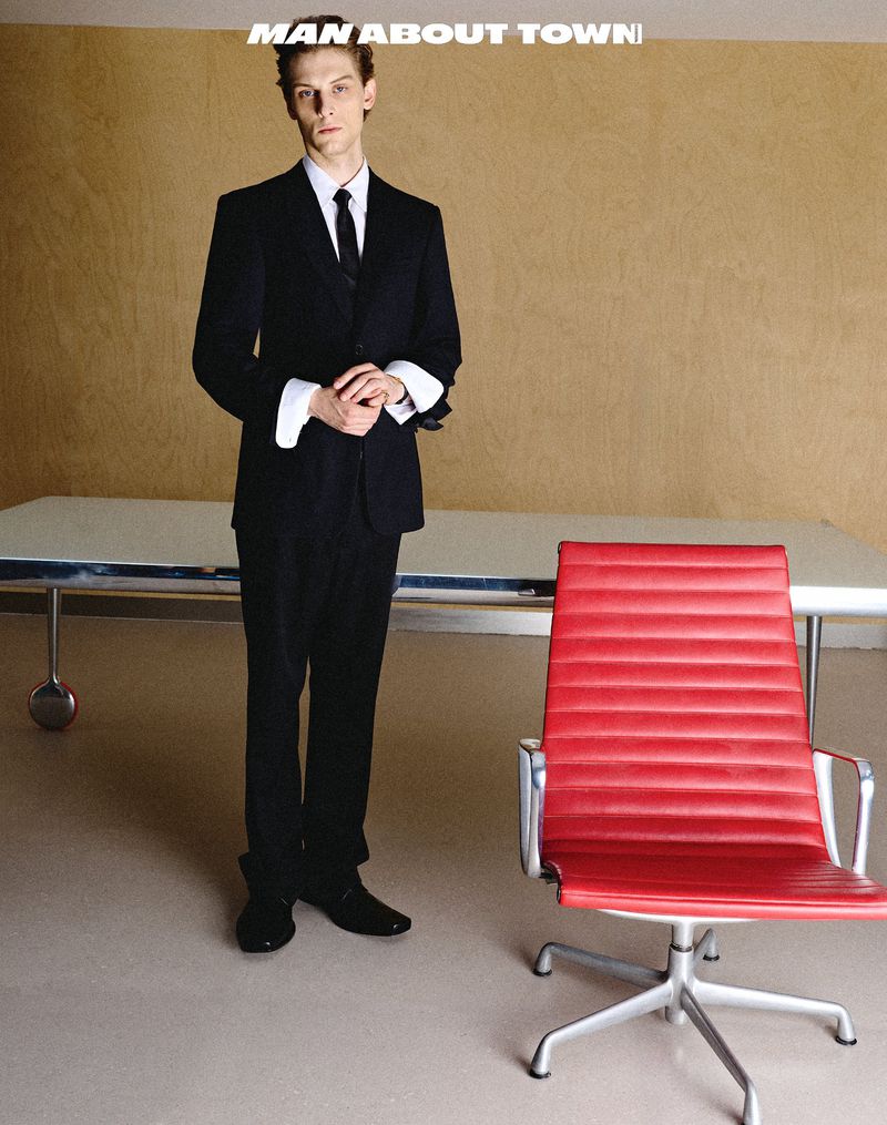 Model wearing a black suit with folded white cuffs stands beside a bright red office chair in a minimalist room.