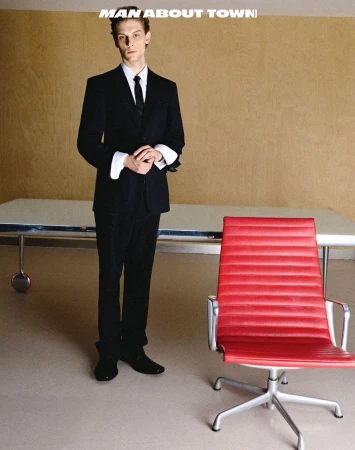 Model wearing a black suit with folded white cuffs stands beside a bright red office chair in a minimalist room.