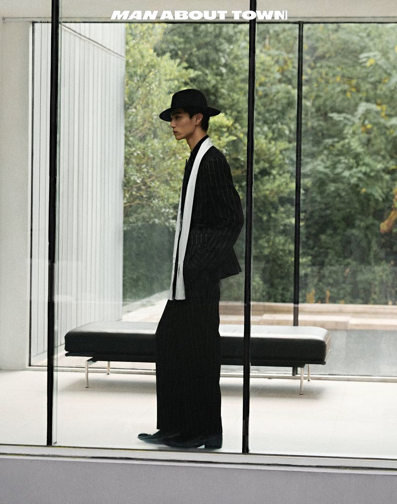 An Asian adult male poses in black pinstripe suit with white scarf and black hat inside a modern glass-walled building with trees outside.