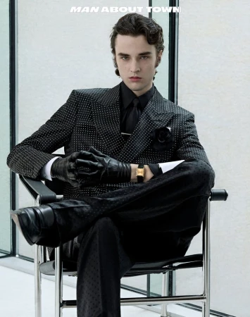 A young adult male with dark hair poses in a patterned black suit, leather gloves, and watch while seated indoors in a minimalist setting.