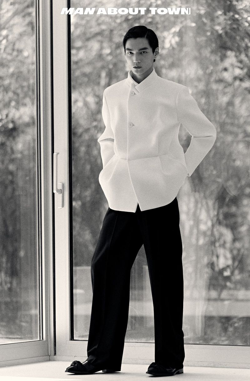 Black and white portrait of an Asian adult male model in a white jacket and black pants, standing near a glass wall with text overlay.