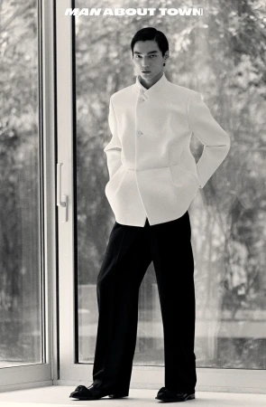Black and white portrait of an Asian adult male model in a white jacket and black pants, standing near a glass wall with text overlay.