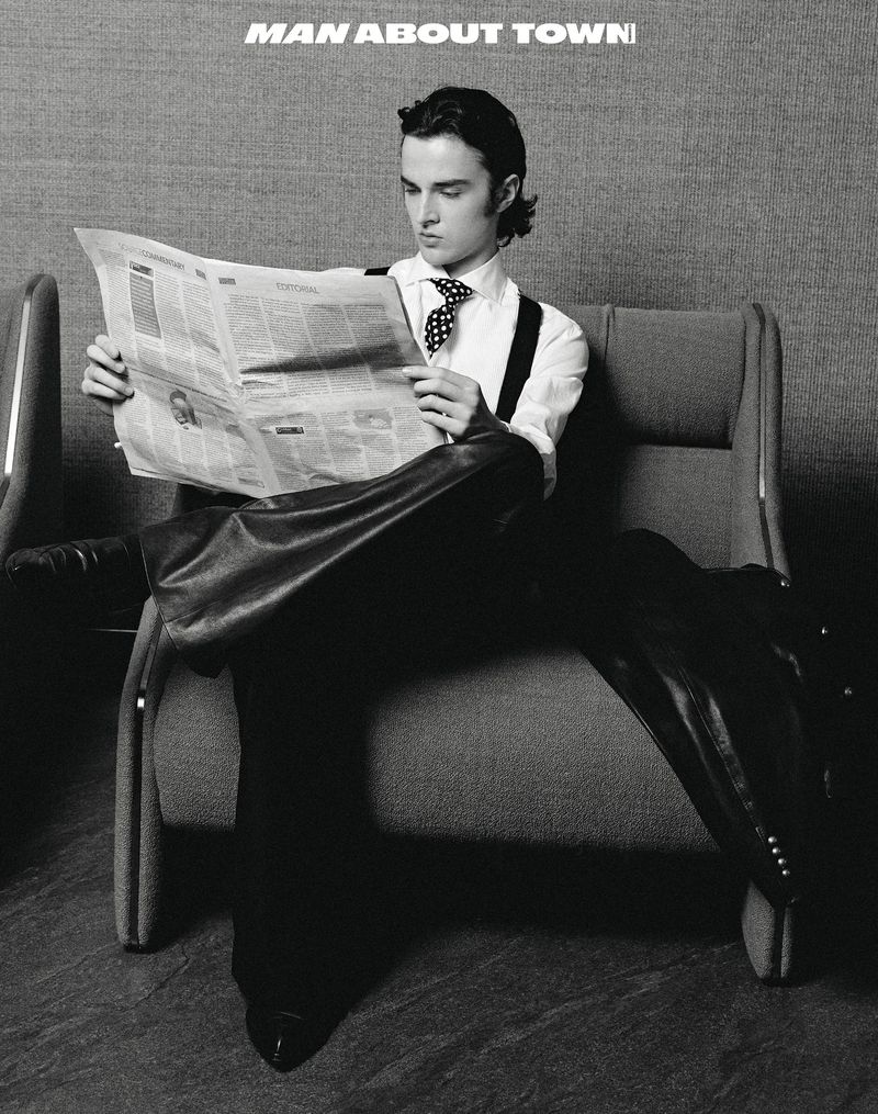 Black and white portrait of a young man in a tie and suspenders sitting on a gray chair reading a newspaper in a minimalist setting.