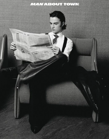 Black and white portrait of a young man in a tie and suspenders sitting on a gray chair reading a newspaper in a minimalist setting.