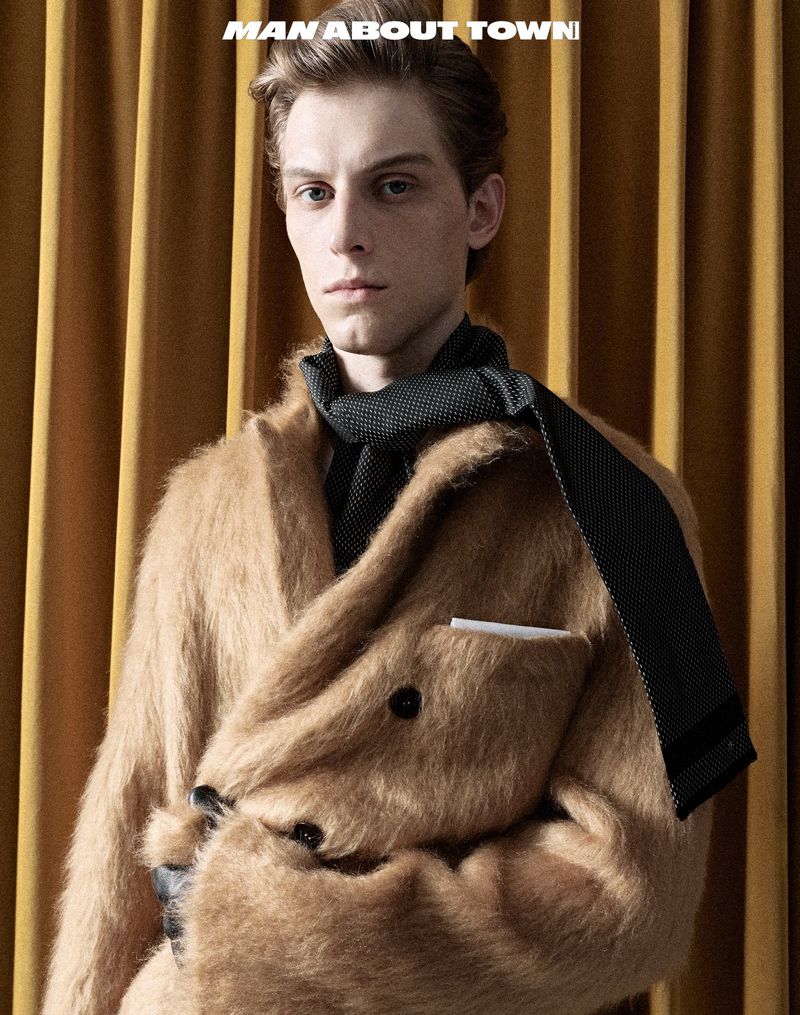 Portrait of a stylish adult male wearing a mohair coat and scarf, posing against a gold curtain backdrop in a studio fashion shot.
