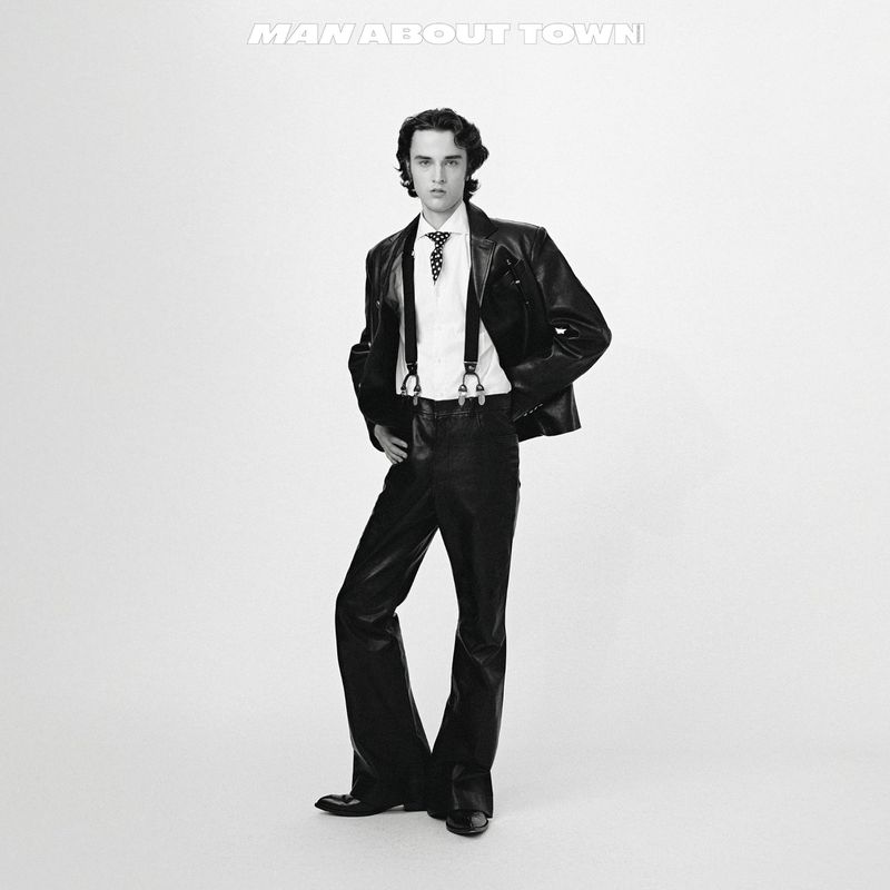 Black and white shot of a man in a leather jacket, dress shirt, and suspenders posing against a white background.