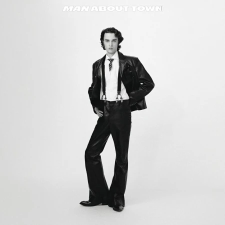 Black and white shot of a man in a leather jacket, dress shirt, and suspenders posing against a white background.