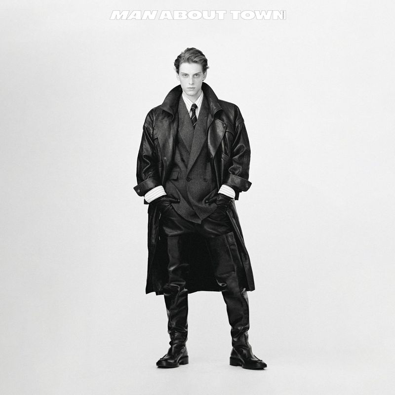 Monochrome portrait of a male fashion model in a full-length pose wearing a dark suit, long leather coat, and boots against a bright white backdrop.