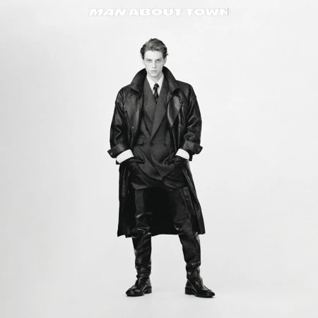Monochrome portrait of a male fashion model in a full-length pose wearing a dark suit, long leather coat, and boots against a bright white backdrop.