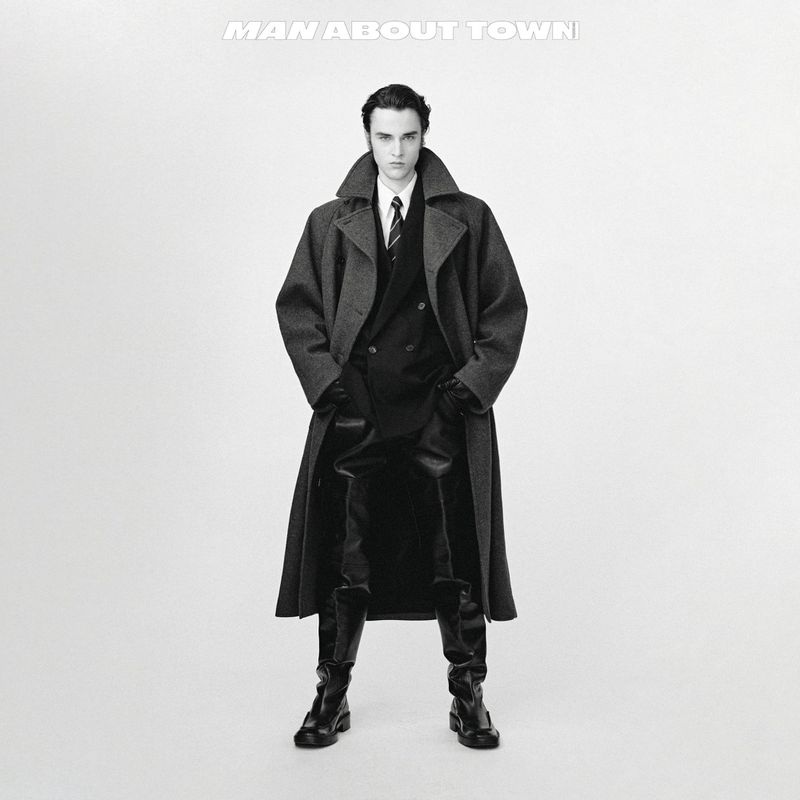 Model wearing an oversized charcoal wool coat, tailored suit, and tall black leather boots against a bright white seamless background.