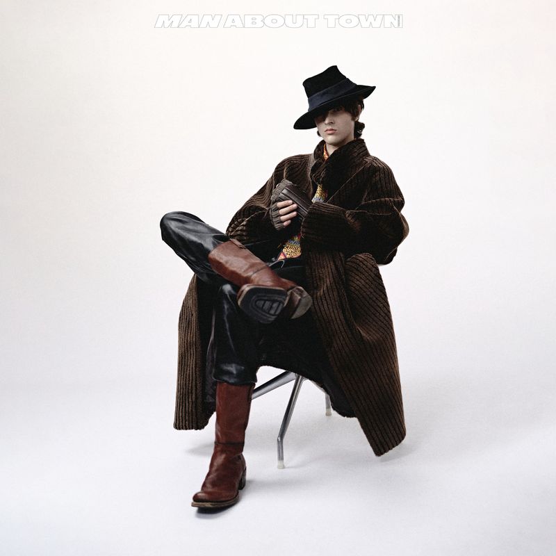 Man in hat and coat sits on a chair, wearing boots, leather pants, and gloves against a plain white background in a studio shot.