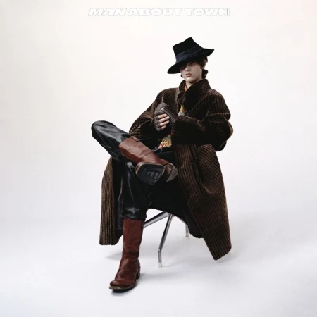 Man in hat and coat sits on a chair, wearing boots, leather pants, and gloves against a plain white background in a studio shot.