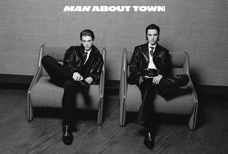 Two adult males sit in modern chairs wearing dark suits and leather jackets against a gray background with the words 'MAN ABOUT TOWN' above them.