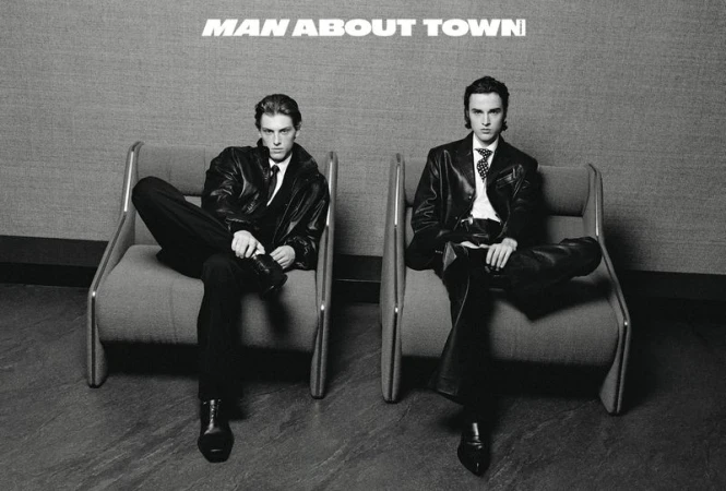 Two adult males sit in modern chairs wearing dark suits and leather jackets against a gray background with the words 'MAN ABOUT TOWN' above them.