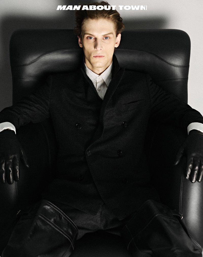 Elegant adult man in a double-breasted suit with leather gloves, poses in a dark chair against a neutral background, fashion editorial shot.