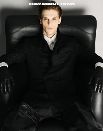 Elegant adult man in a double-breasted suit with leather gloves, poses in a dark chair against a neutral background, fashion editorial shot.