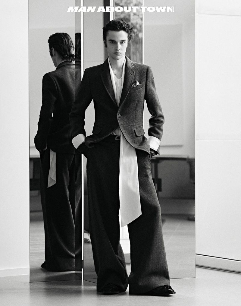 Monochrome fashion shot of a slender man in a dark suit and wide-leg pants posing in front of a mirror, reflecting his back.