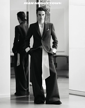 Monochrome fashion shot of a slender man in a dark suit and wide-leg pants posing in front of a mirror, reflecting his back.