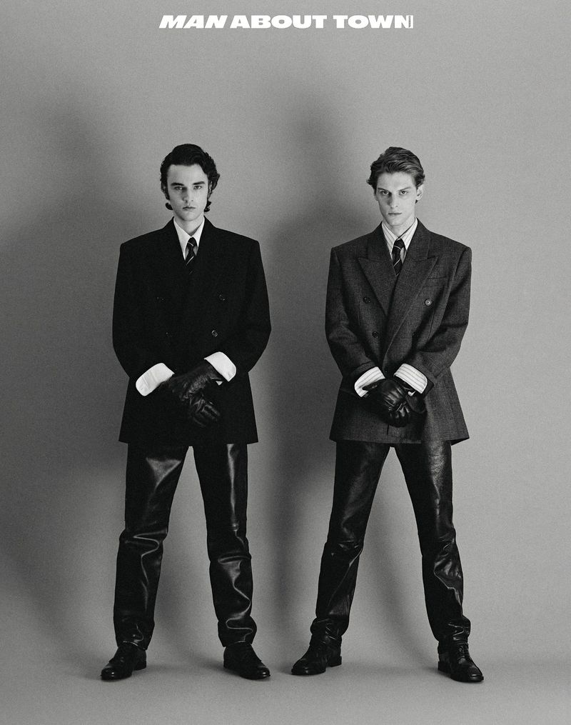 Two stylish adult men in tailored suits and leather gloves pose indoors against a grey backdrop for a fashion editorial.