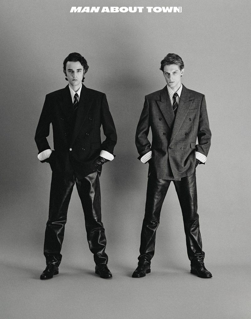 Two young men pose in suits and shiny pants against a grey backdrop, editorial fashion with a vintage aesthetic. 