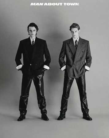 Two young men pose in suits and shiny pants against a grey backdrop, editorial fashion with a vintage aesthetic. 