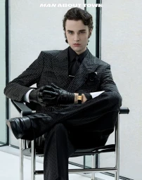 A seated male model wearing a black patterned suit, black shirt, and leather gloves poses in a modern interior.