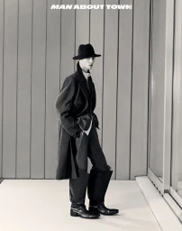East Asian model in a black fedora, long dark coat, and structured leather harness boots posed against vertical wall paneling.