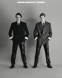 Two male models posing in tailored double-breasted blazers, neckties, and black leather trousers against a seamless grey backdrop.