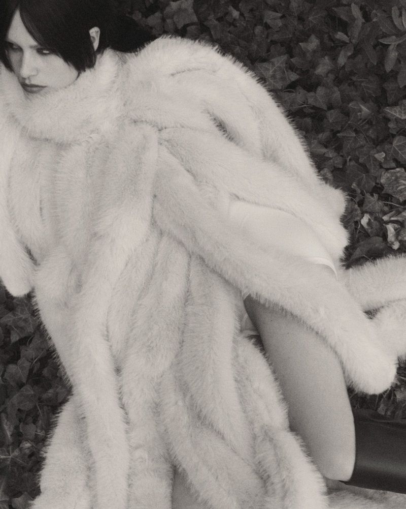 Model Merel Roggeveen reclines in an oversized white fur coat surrounded by dark ivy leaves.