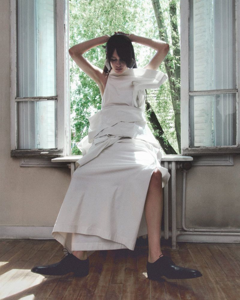 Fashionable woman in a white dress sitting on a window sill with her hands up, trees visible outside the window, indoor, medium shot.