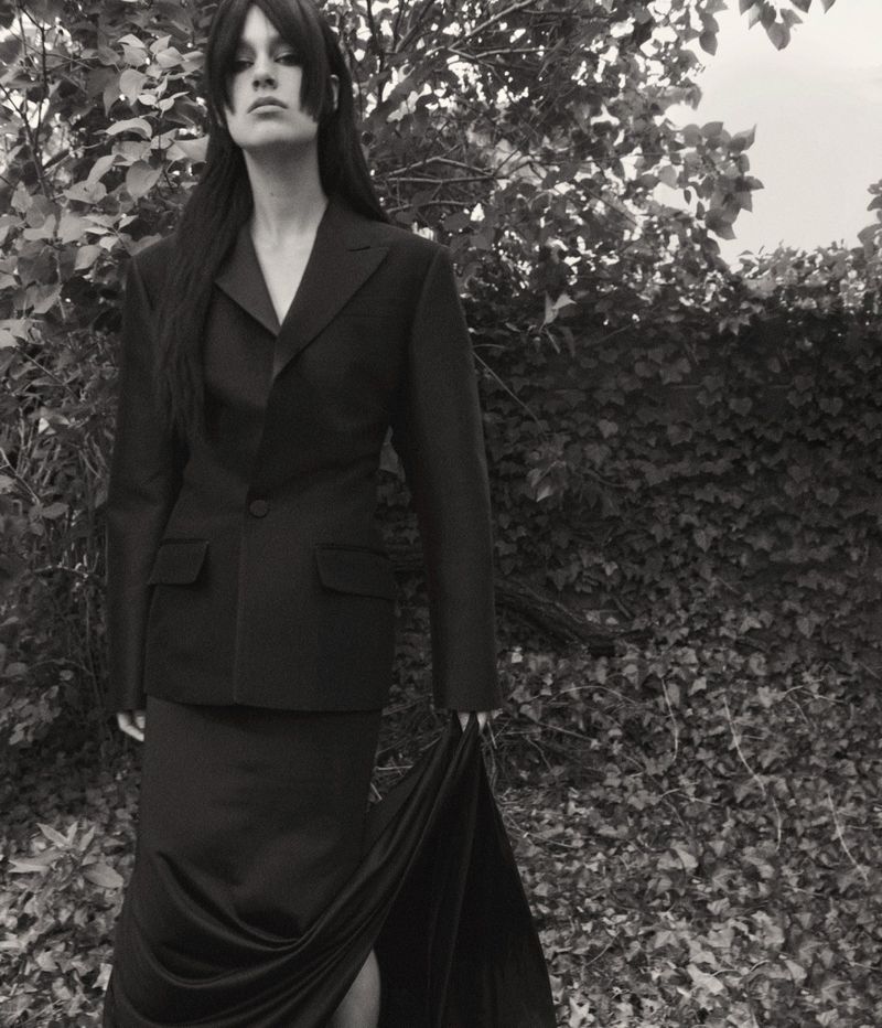 A woman in a black suit and long skirt stands in an outdoor setting with leafy trees. The image is black and white.