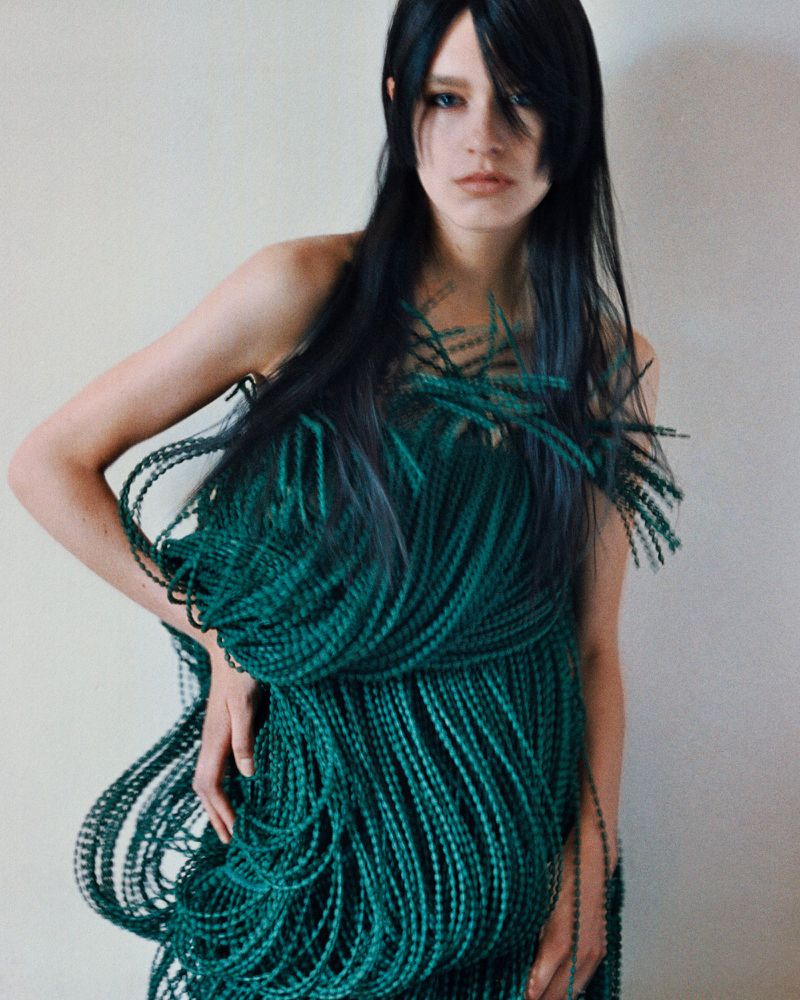 Model Merel Roggeveen wears a sculptural emerald green braided cord dress against a plain neutral studio background.