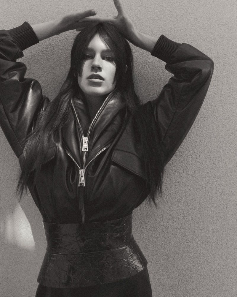 A slender woman with long brown hair poses in a black leather jacket and belt, hands raised above her head against a gray background.