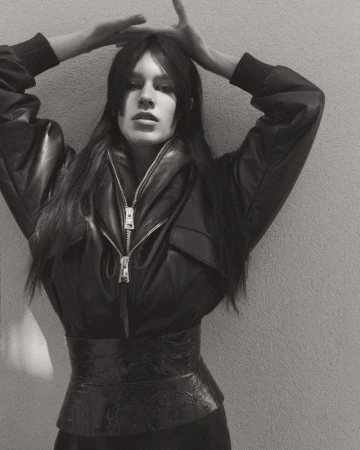A slender woman with long brown hair poses in a black leather jacket and belt, hands raised above her head against a gray background.