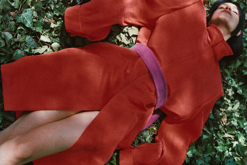 Woman in a red outfit lies on a bed of ivy, relaxed with eyes closed, in outdoor setting with soft natural light, fashion editorial photography.