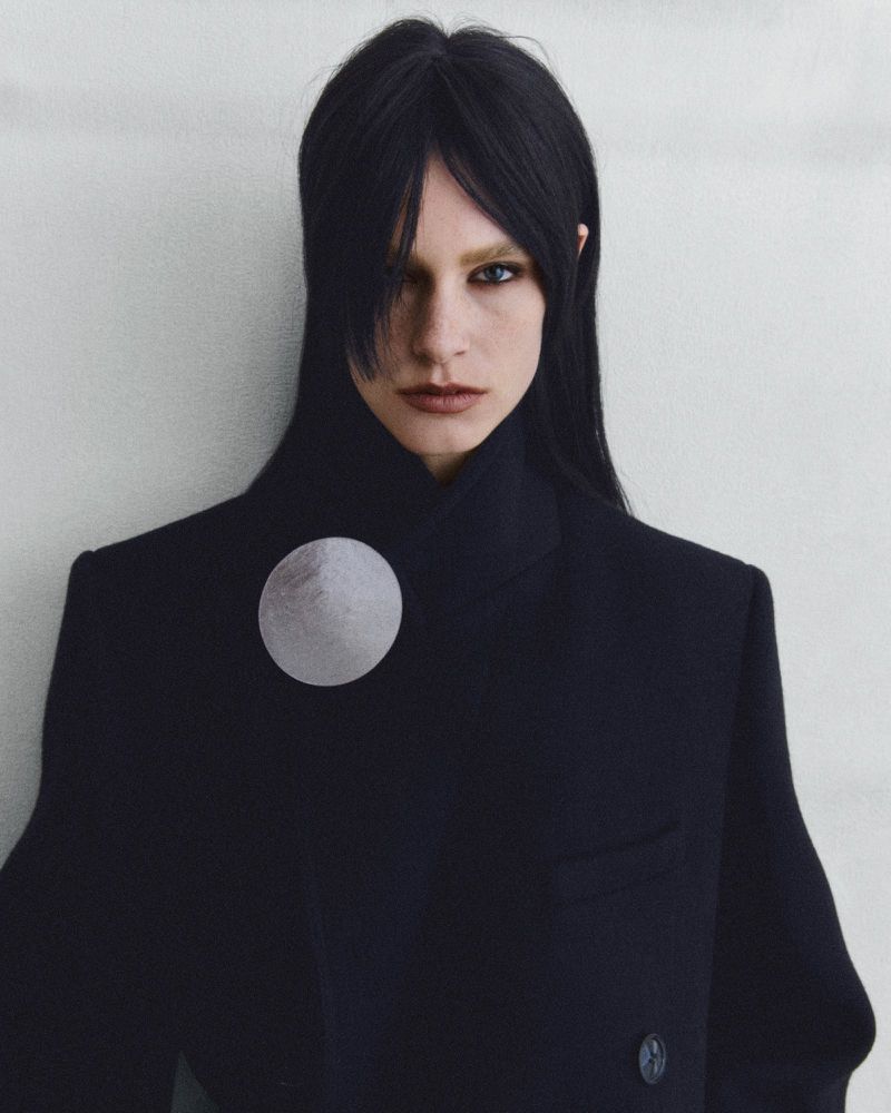 Portrait of a woman with black hair wearing a high-collared dark jacket with a silver moon brooch against a muted background.