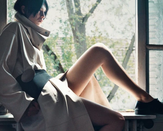 Brunette woman in a beige trench coat poses in a window with a tree and building visible outside, showcasing a fashion editorial look.