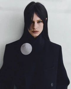 Model Merel Roggeveen wearing a tailored black coat with a circular silver brooch against a neutral studio backdrop.