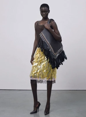 Model wearing a yellow sequined skirt and carrying an oversized black feathered shoulder bag against a light studio backdrop.