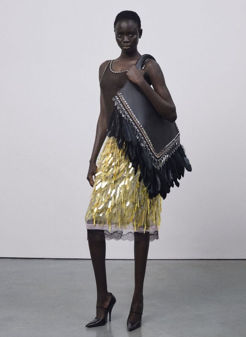 Fashion shot of a black female model wearing a brown top, gold fringe skirt, black high heels, and carrying a feather-trimmed bag in studio.