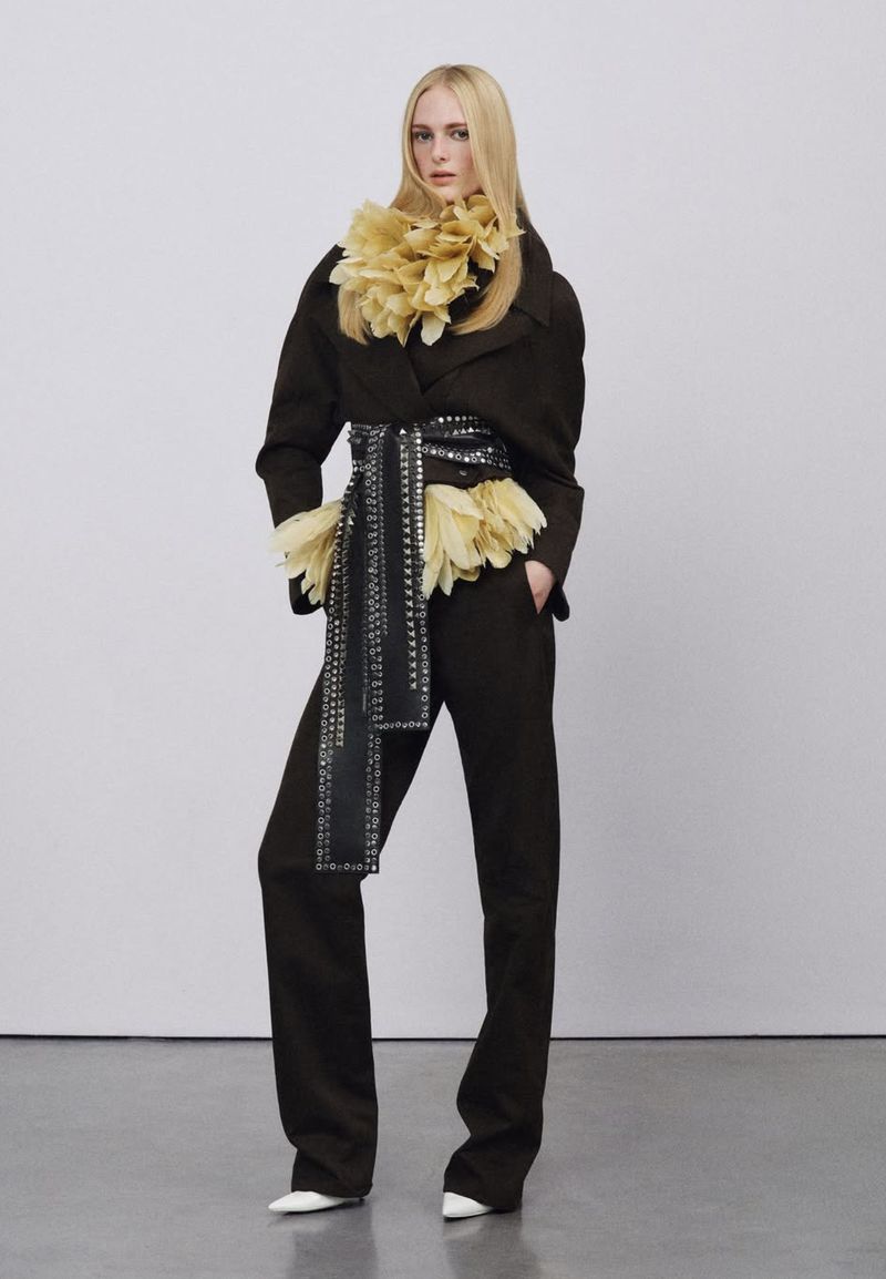 A full shot of a blonde model wearing a brown jacket and pantsuit, accentuated with a textured belt and neck piece, against a grey studio backdrop.