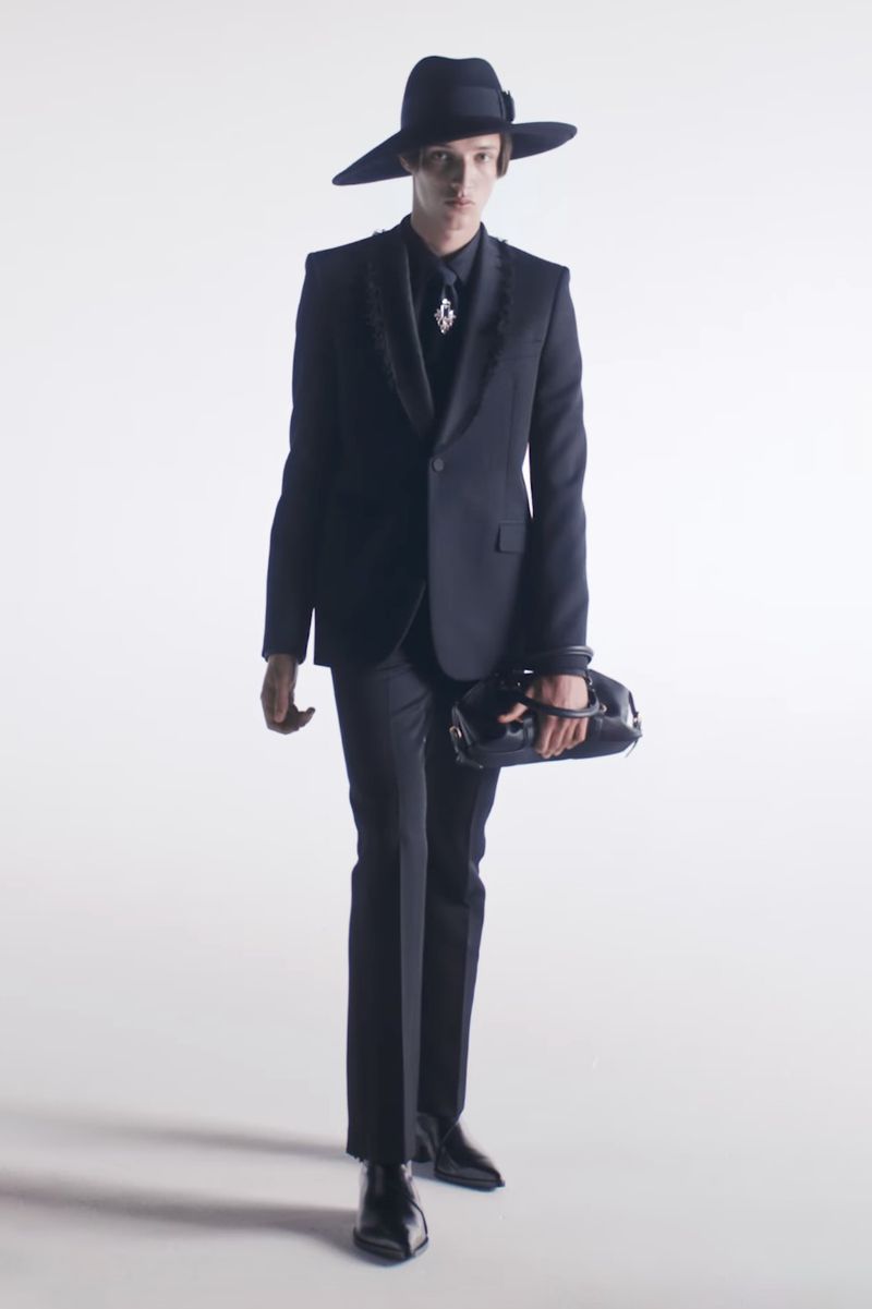 Full-length shot of adult male wearing black suit and wide-brim hat, carrying purse, posing against white background in a fashion editorial.