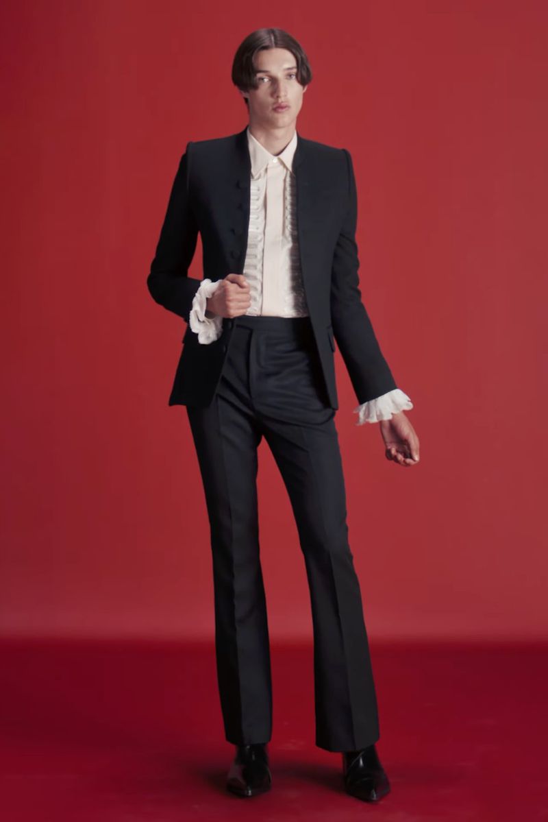 Portrait of a young adult man in a dark suit and frilled shirt, standing against a red backdrop in a fashion-forward studio shot.