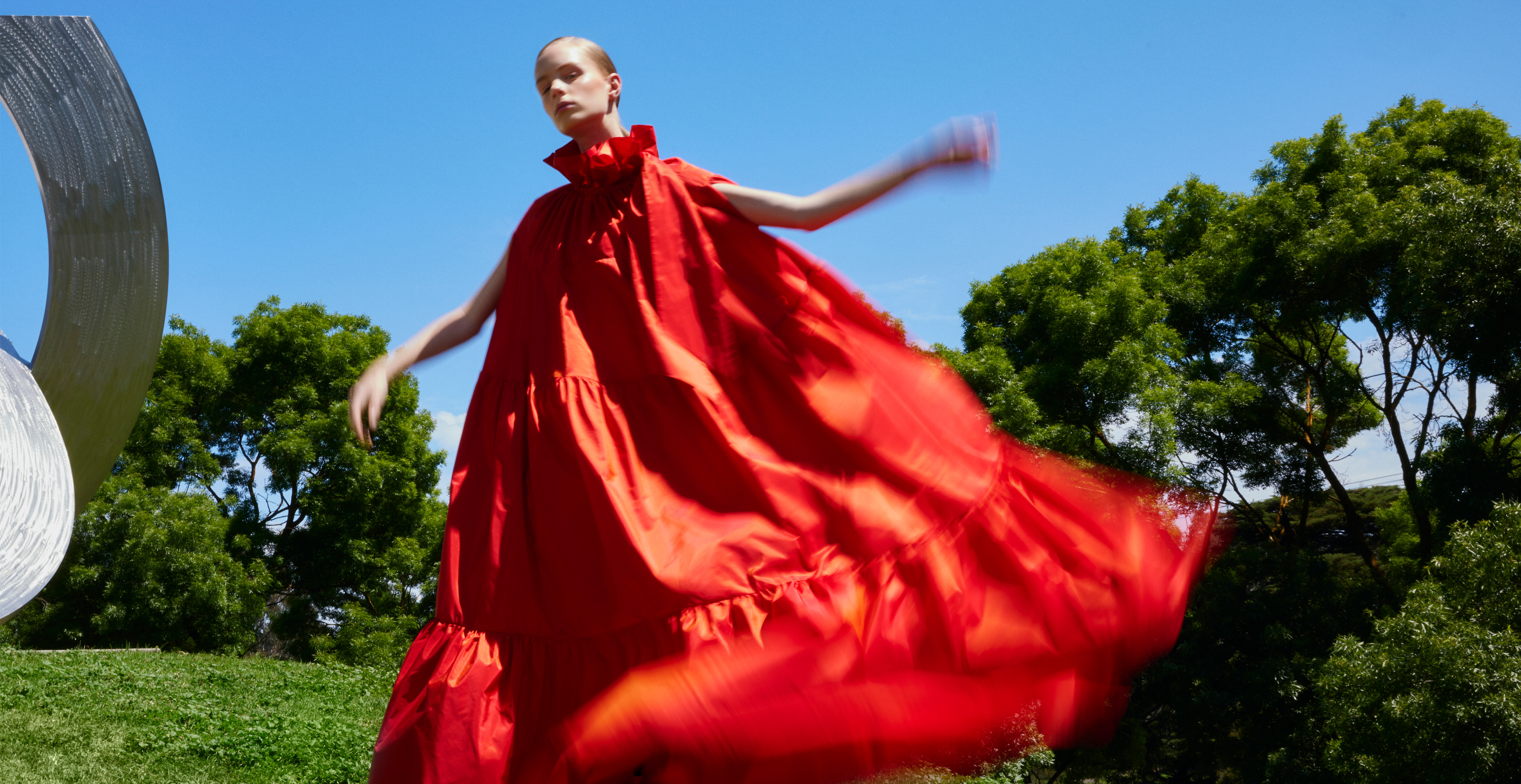 Woman in billowing red dress poses in an outdoor setting with green trees and blue sky, accentuated by a metallic sculpture.