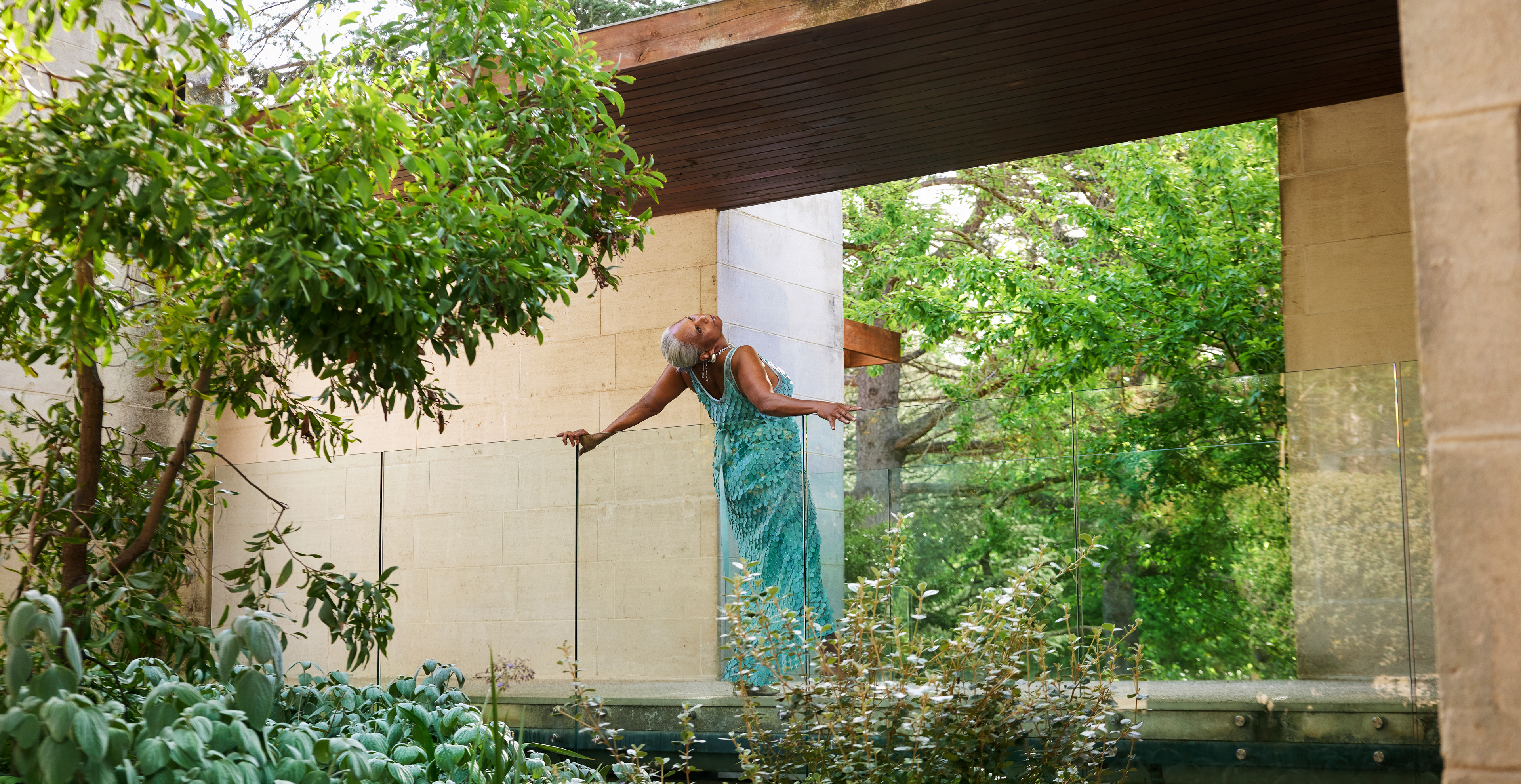 Older woman in teal dress strikes a pose in lush outdoor garden, framed by architectural elements, showcasing natural light and vivid greenery.