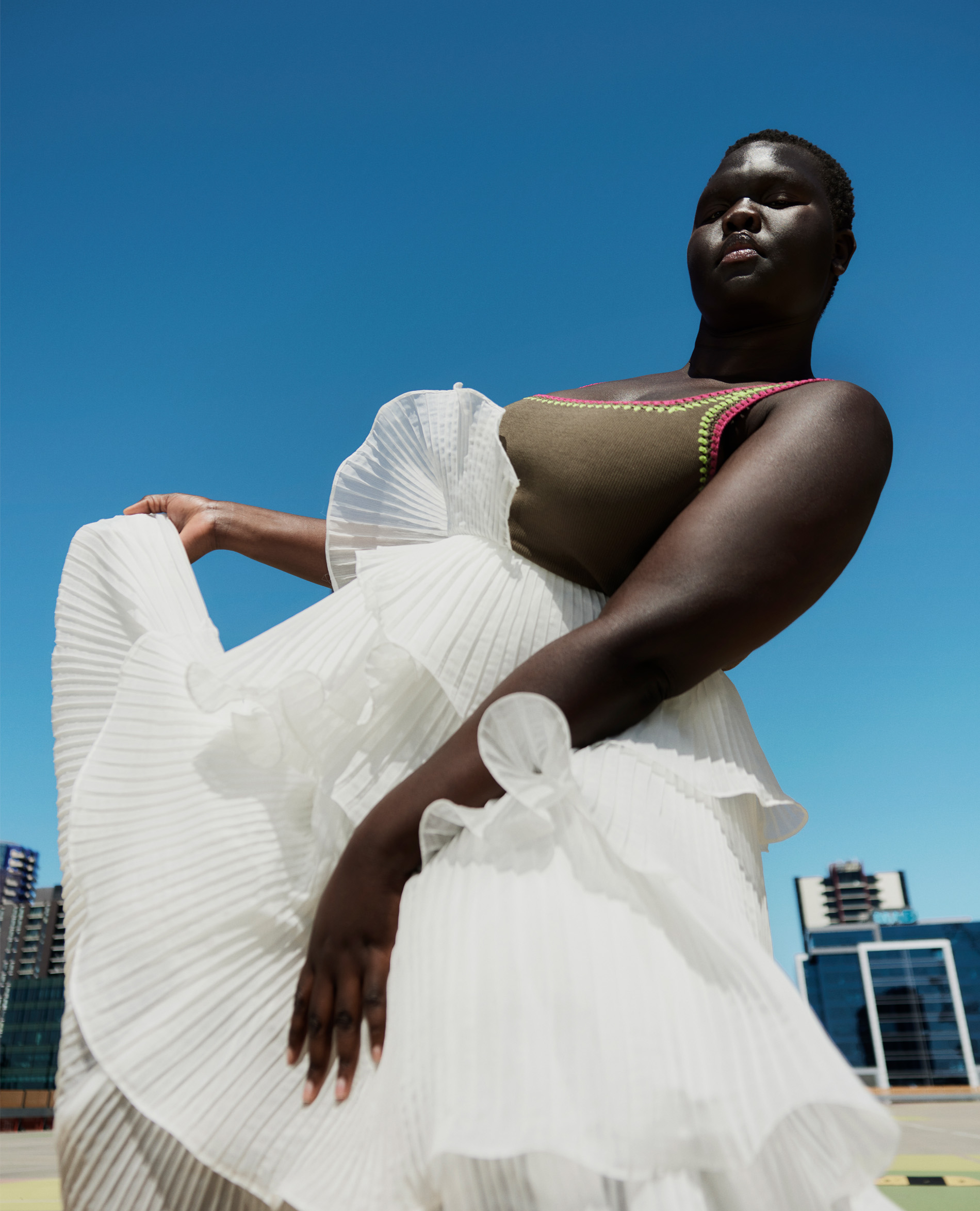Woman in flowing white dress poses against a blue sky, with city buildings visible in the background. Fashionable attire and outdoor setting.