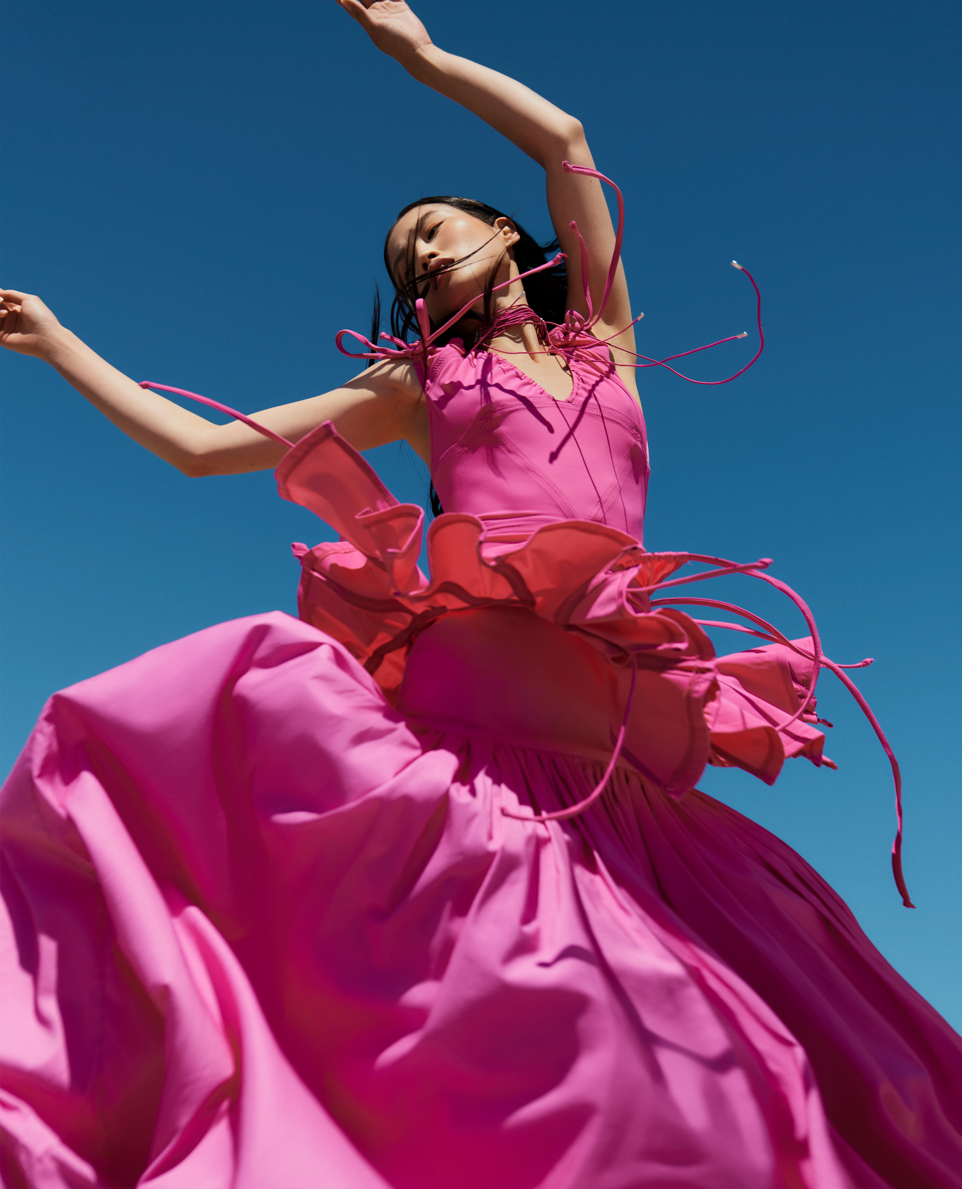 Woman in pink dress with a ruffled waist posing against a bright blue sky, arms raised, low angle, fashion editorial style.