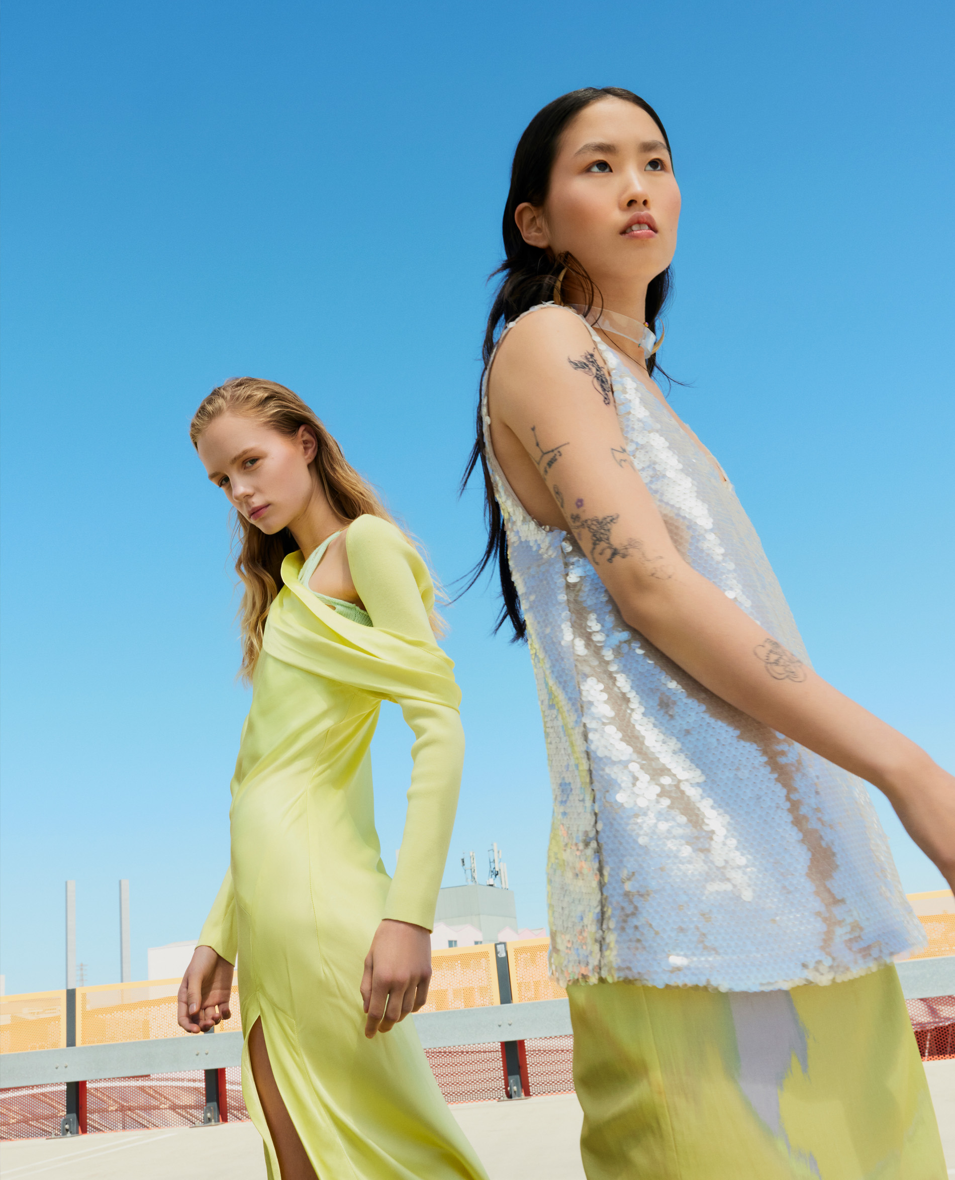 Two women in dresses stand on a rooftop against a blue sky. One wears a yellow dress with a slit, while the other has a silver top.